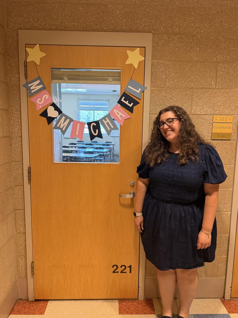 Ellie outside the door to her classroom that is decorated with a banner of her name.